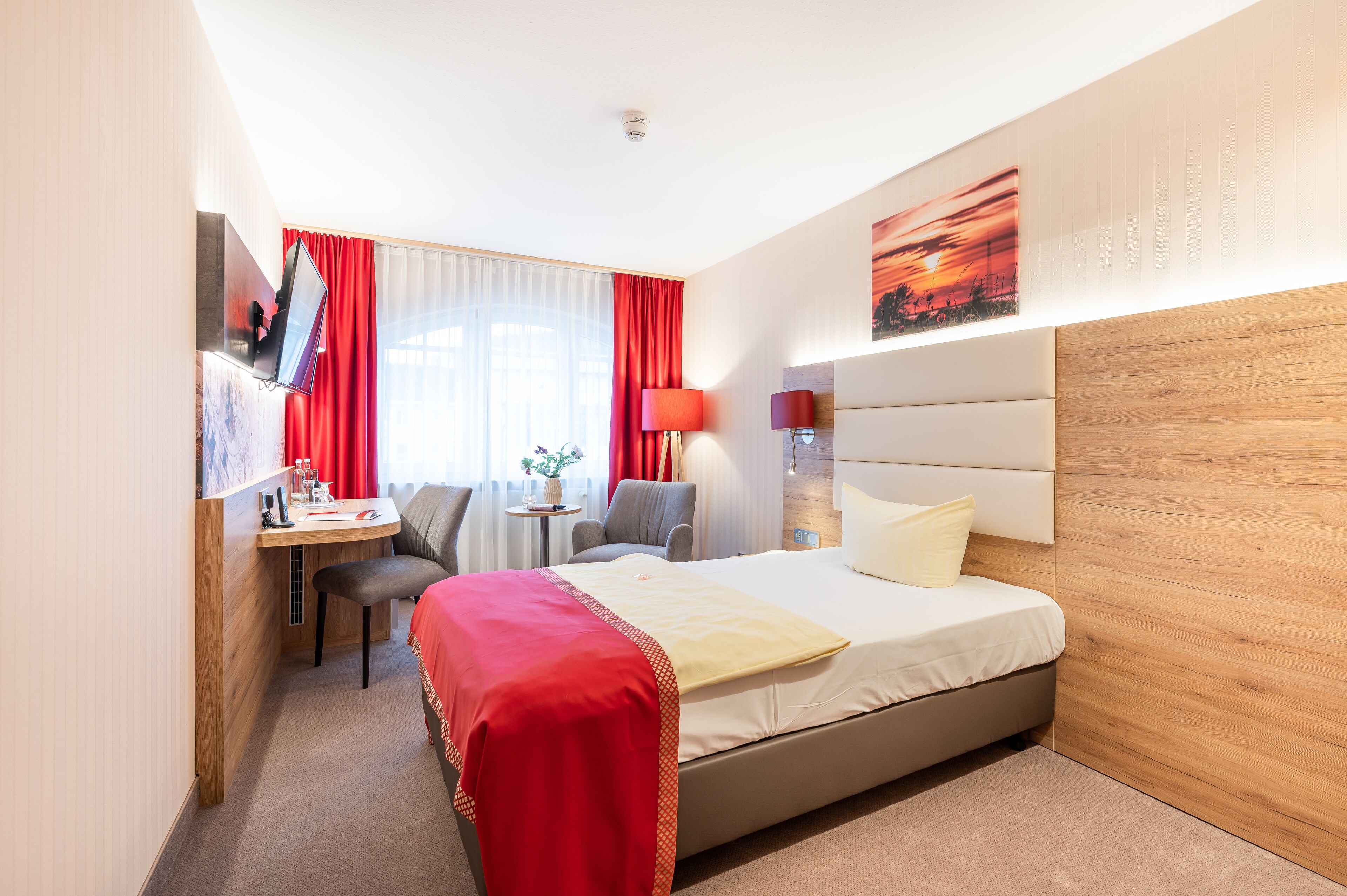 Ett hotellrum med en säng, ett skrivbord, en stol, en tv och ett fönster med röda gardiner.