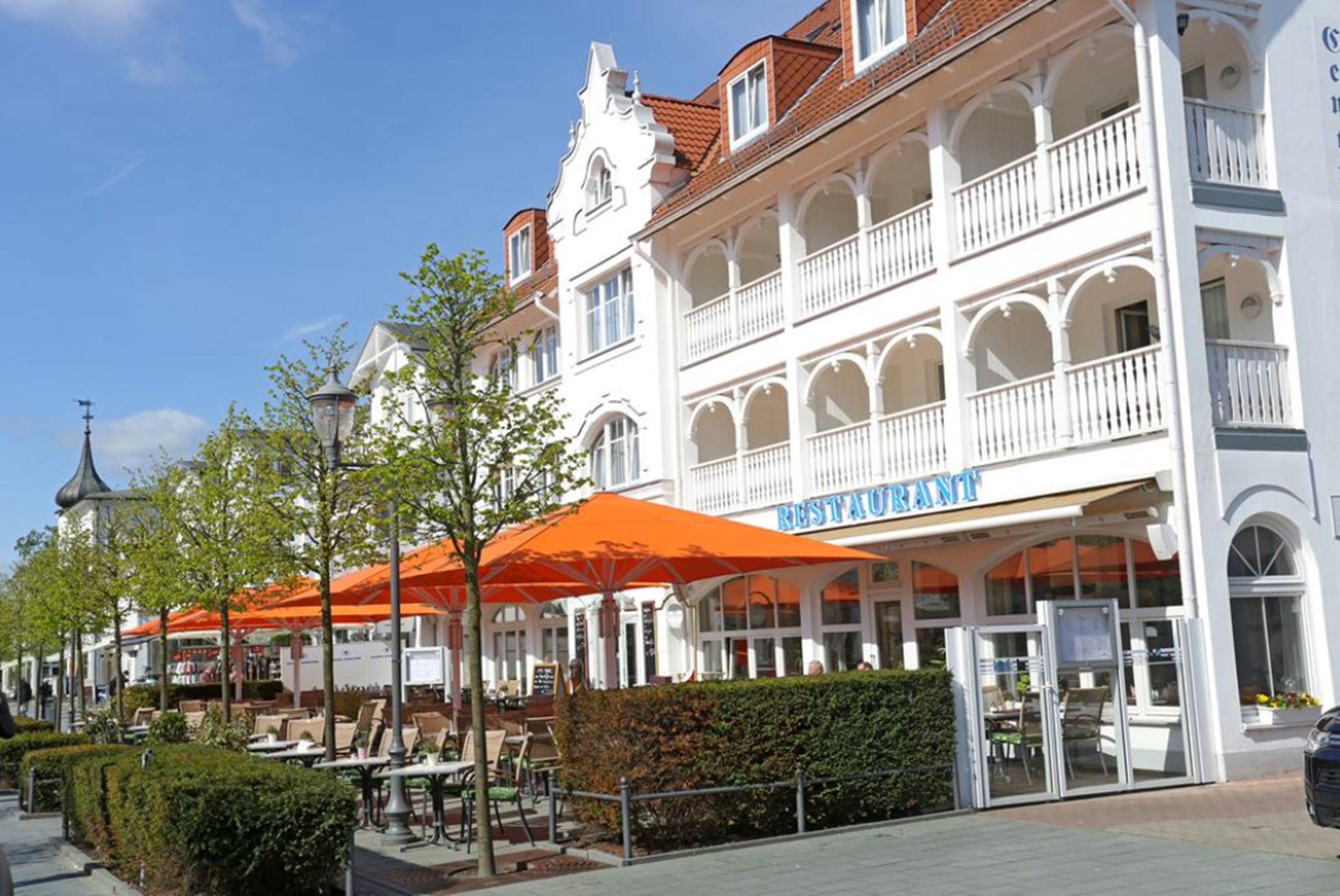 A white building with a restaurant named "RESTAURANT" on the ground floor, outdoor seating with orange umbrellas, and a clear blue sky.