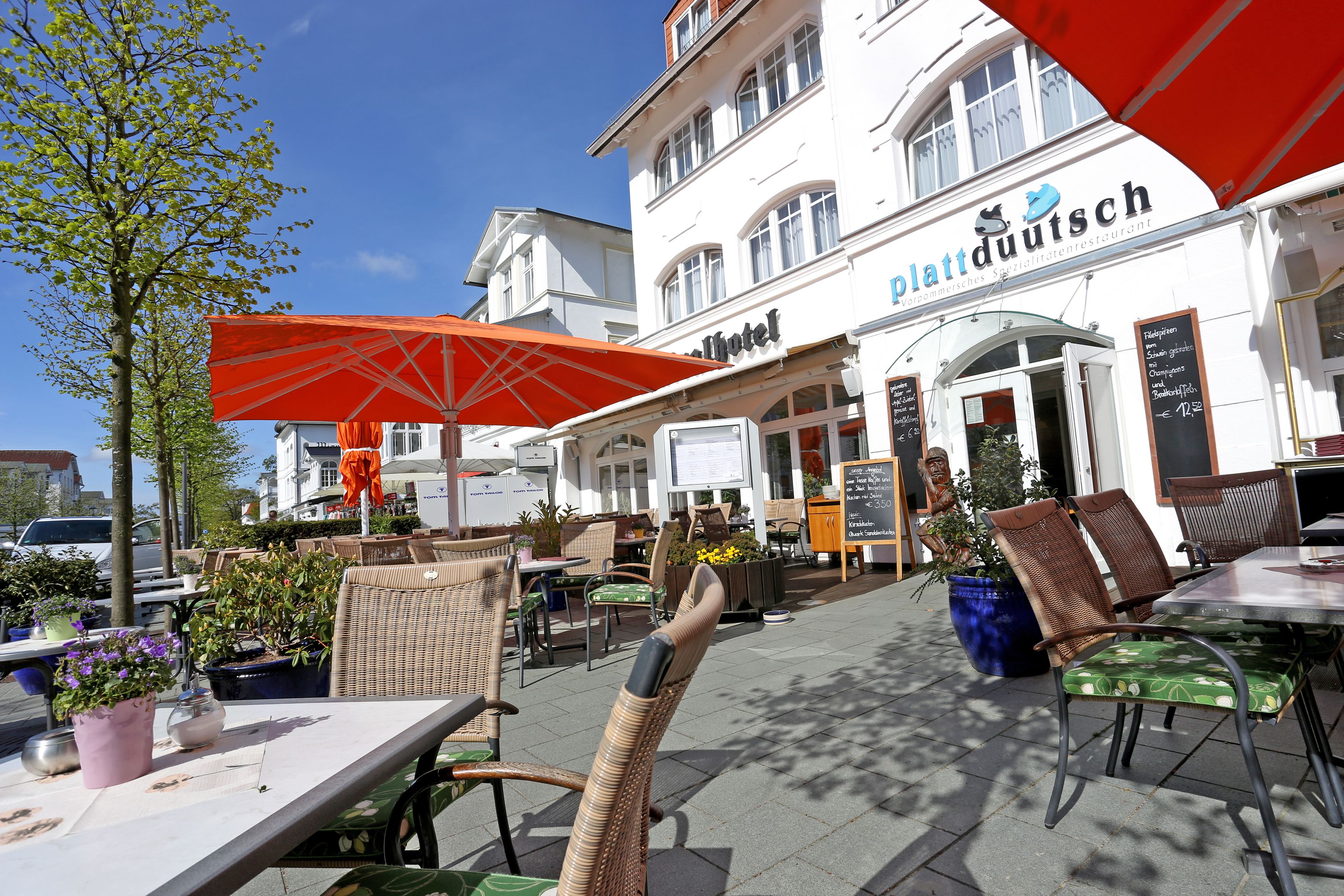 An outdoor cafe area with wicker chairs and tables, a large orange umbrella, and a building with the sign "plattduutsch".
