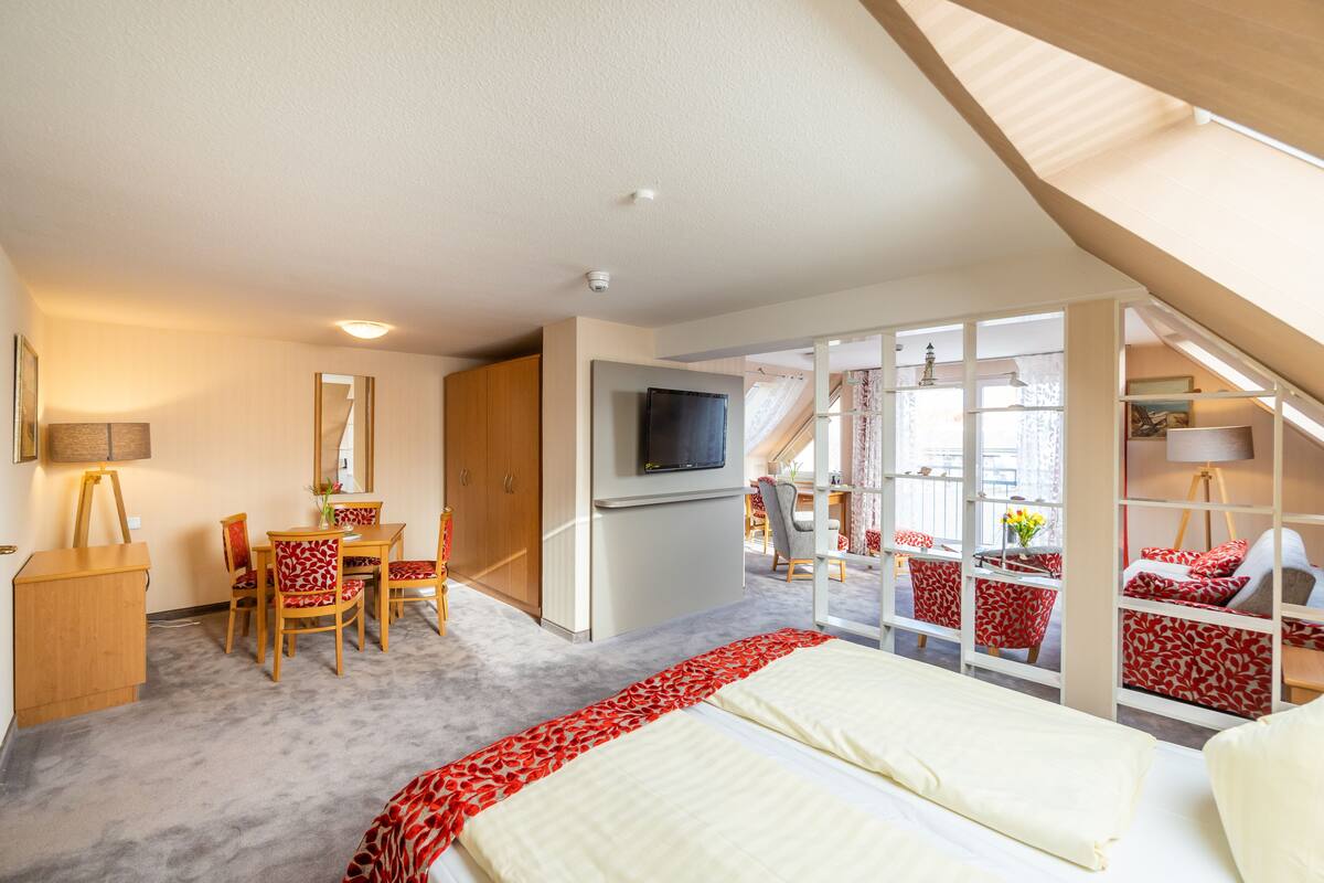 Une chambre à coucher comprenant un lit, un bureau, une chaise, un téléviseur et un balcon doté de meubles aux motifs rouges et blancs.