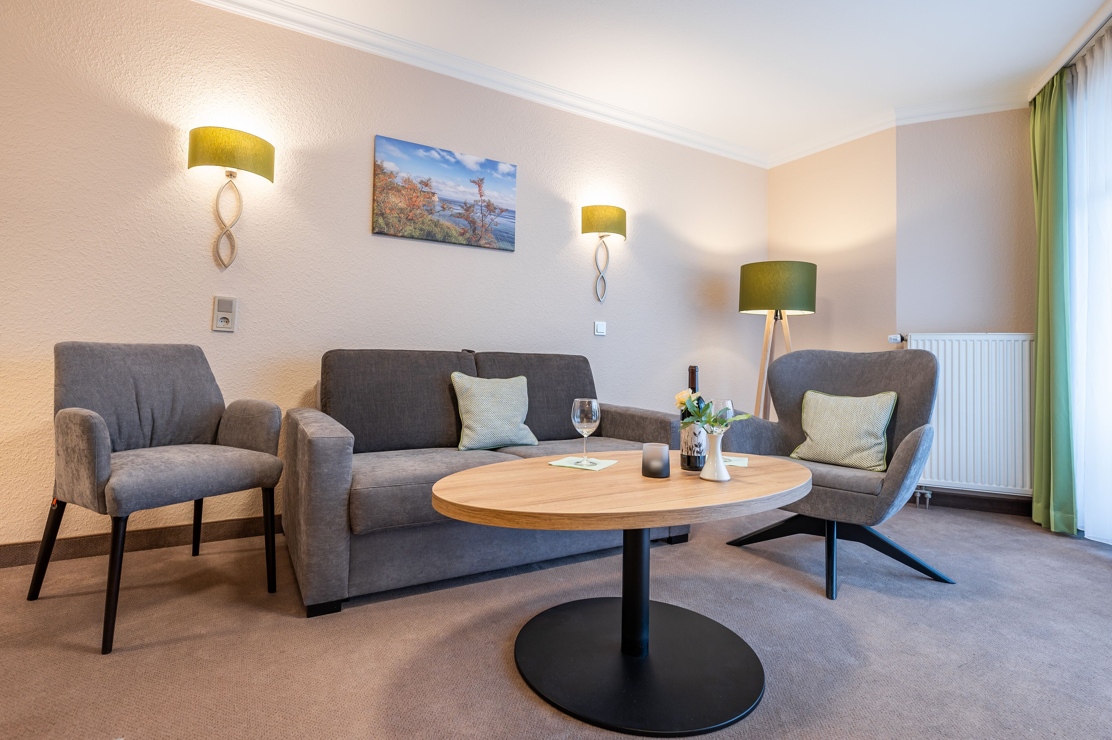 A modern hotel room with a grey sofa, two armchairs, a round wooden table, and wall-mounted lamps.