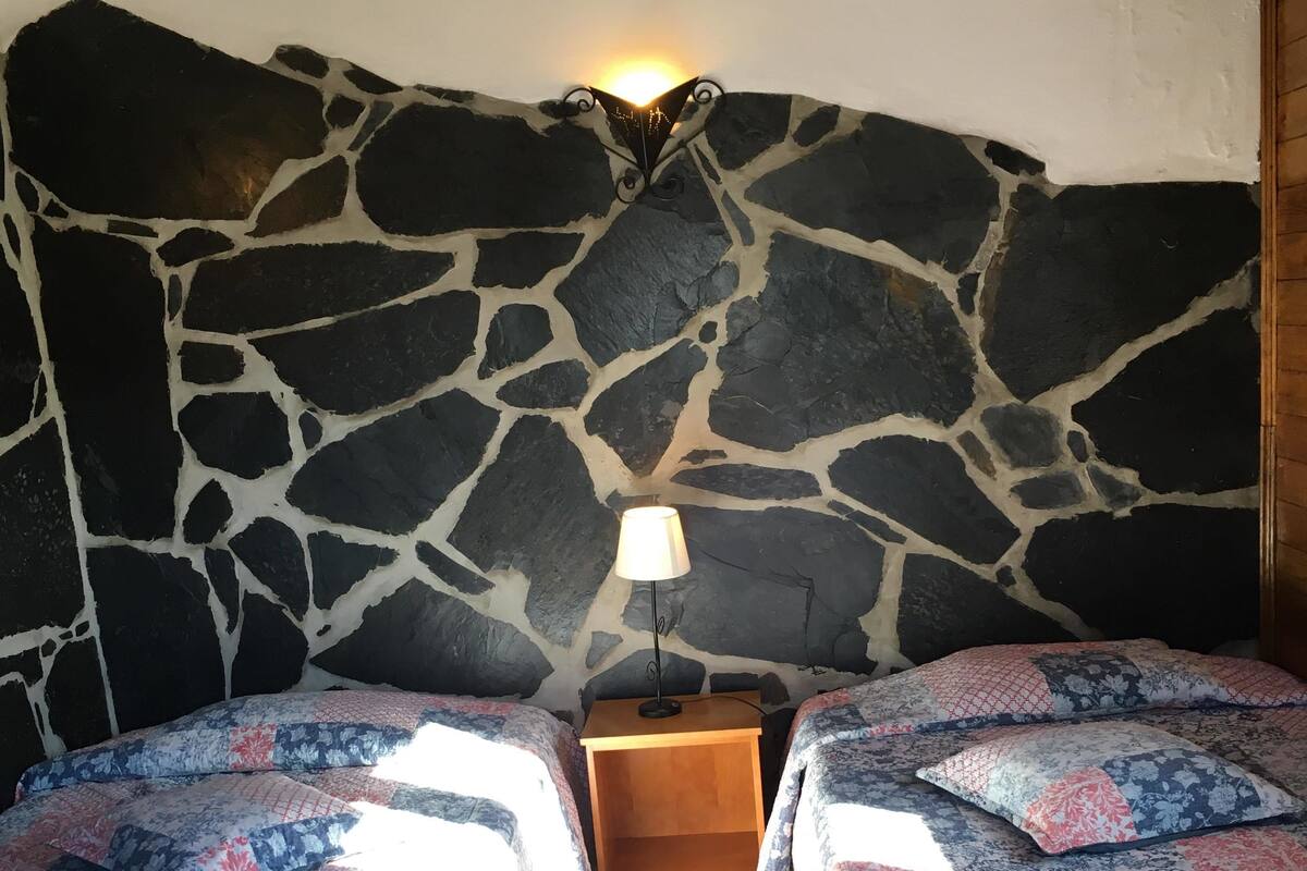 A room with two beds, a stone wall, a nightstand with a lamp, and a wooden floor.