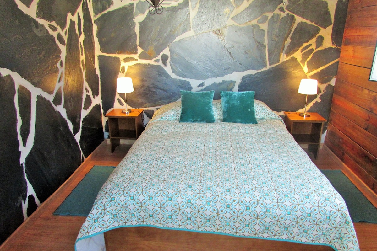 A bed with a patterned bedspread, two pillows, and two bedside lamps in a room with a stone wall.