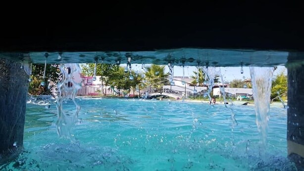A clear view of a swimming pool with water splashing, surrounded by a tropical setting with palm trees and buildings in the background. - Lusitaurus (Redondo)