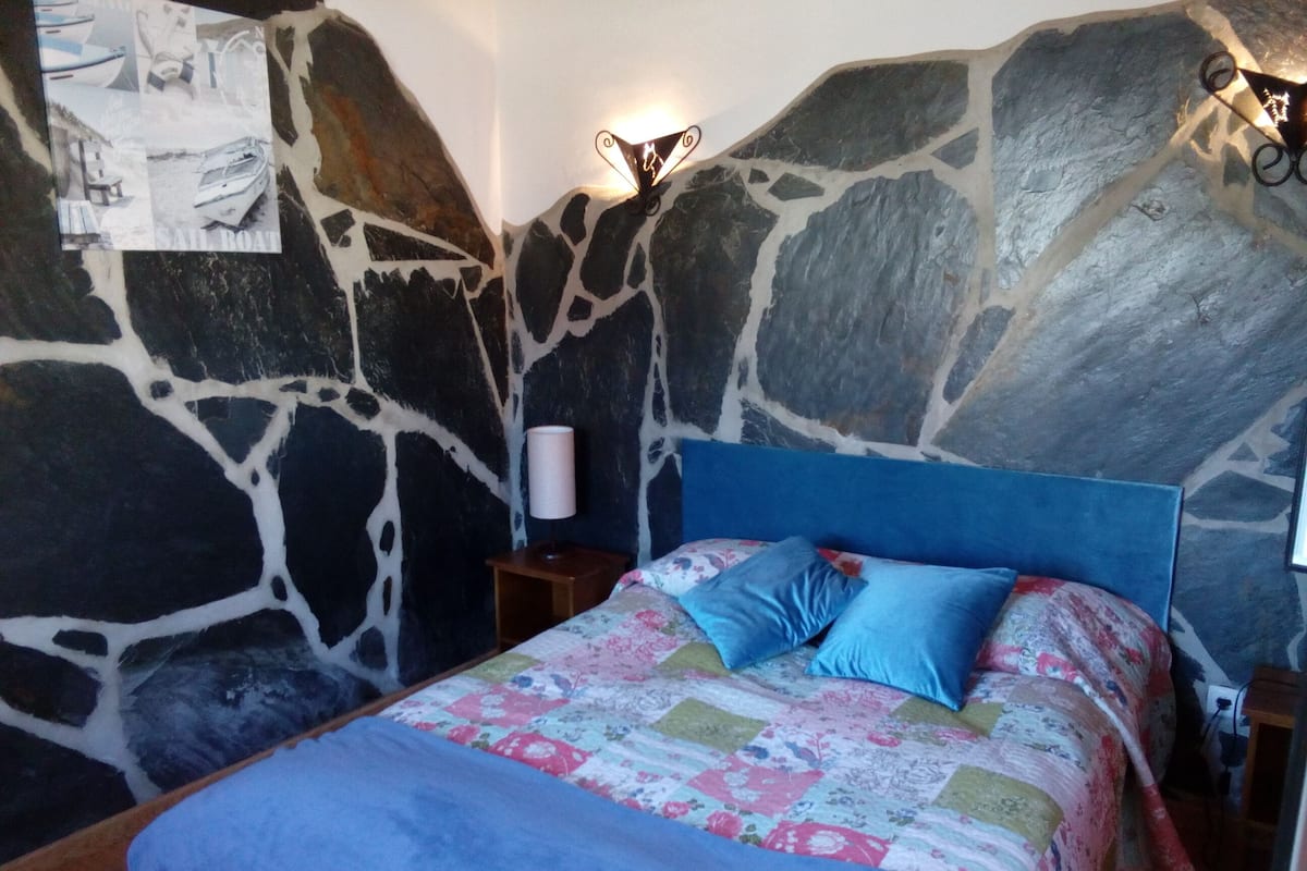A bedroom with a stone wall, a bed with a blue headboard, and a floral bedspread.