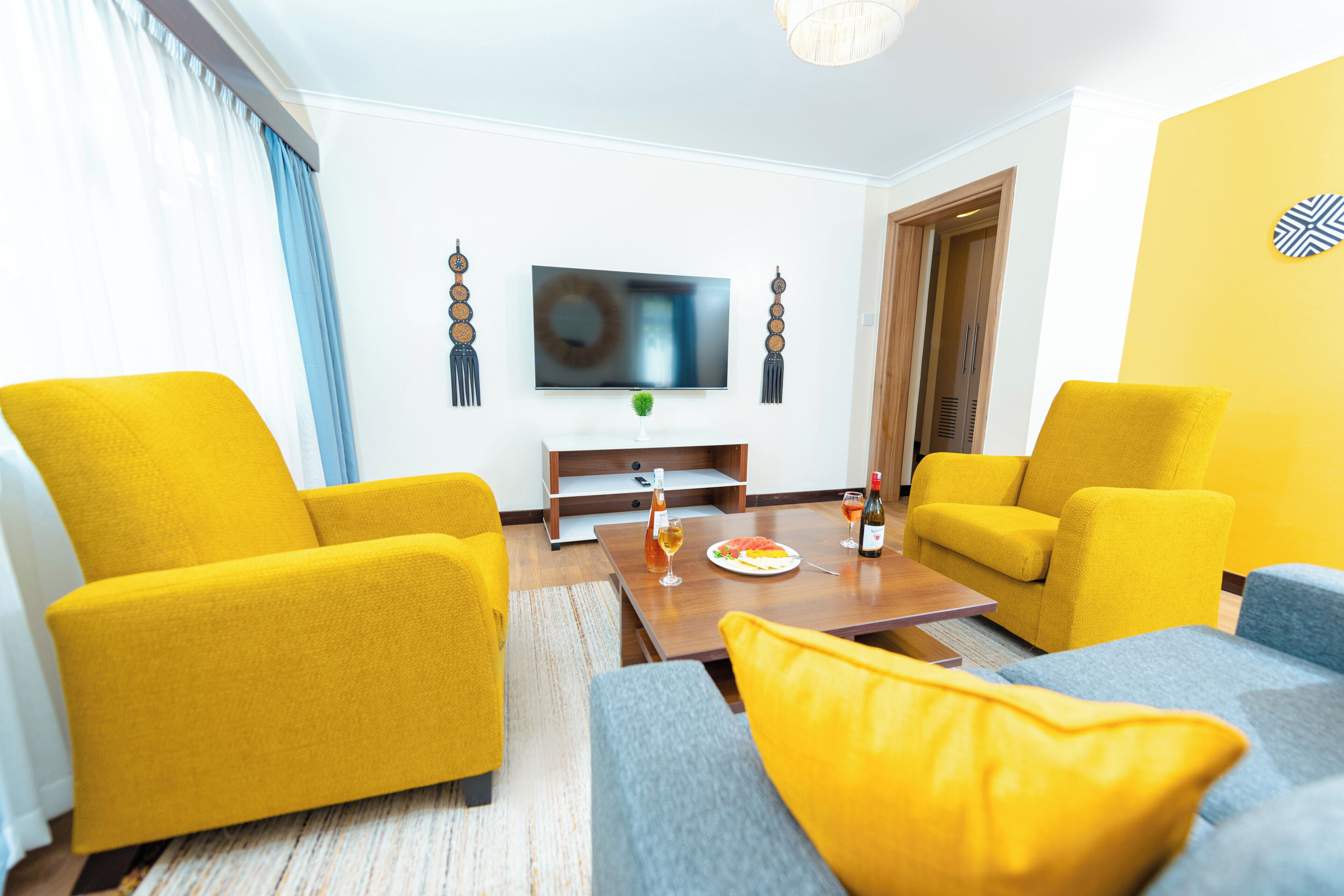 A modern living room with yellow armchairs, a grey sofa, a wooden coffee table, and a flat-screen TV mounted on the wall.