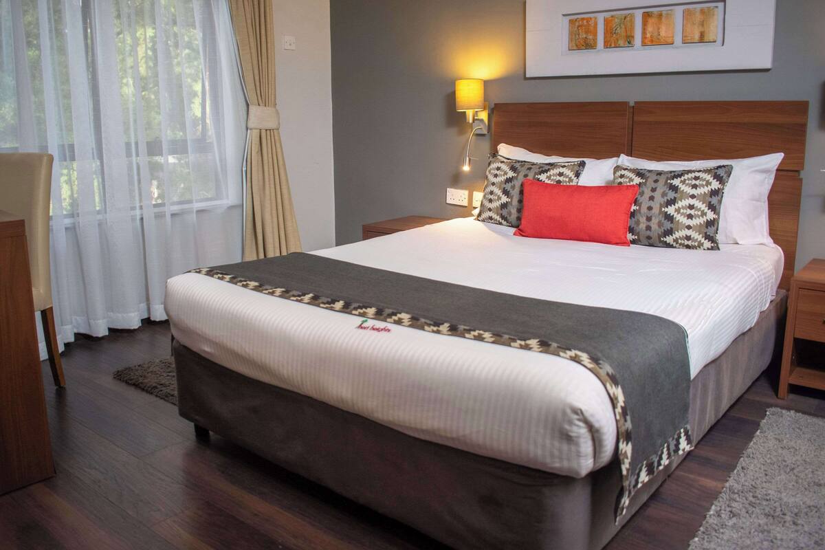 A neatly made bed with a patterned bedspread and pillows, a bedside lamp, and a window with sheer curtains.