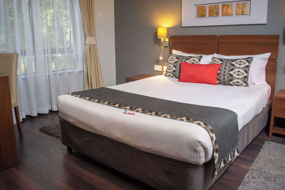 A neatly made bed with a patterned bedspread and pillows, a bedside lamp, and a window with sheer curtains.