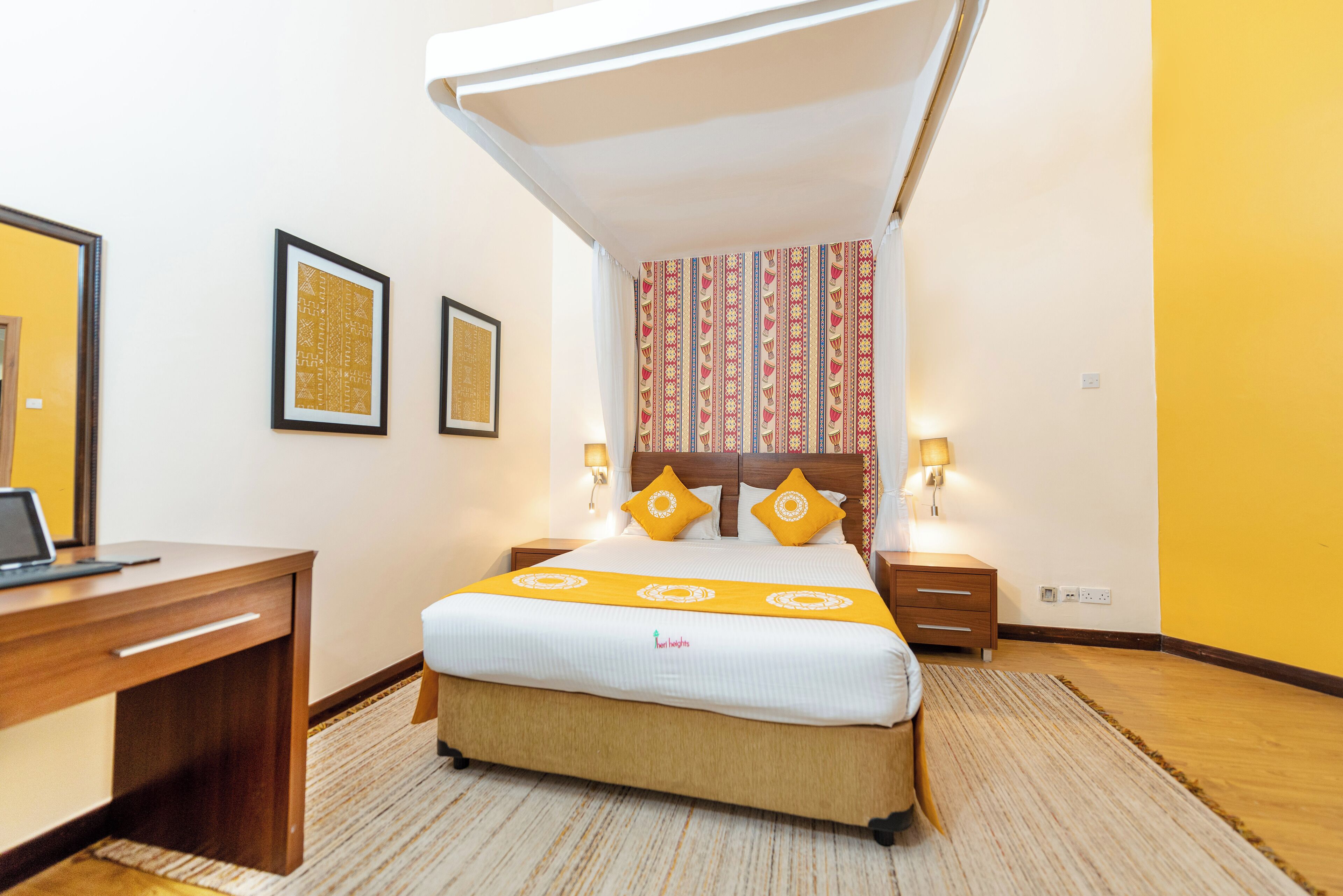 A hotel room with a bed, bedside tables, a desk, and framed pictures on the wall.