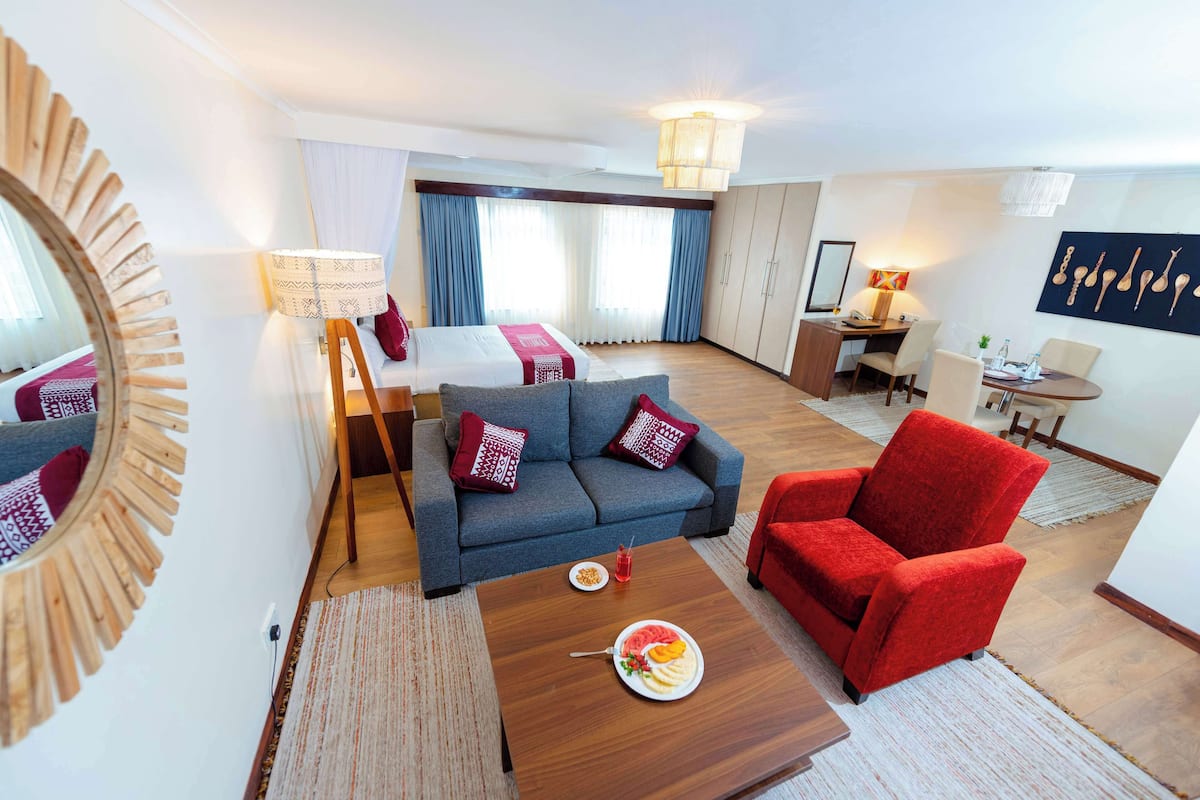A hotel room with a bed, a sofa, a red armchair, a dining table with a plate of food, and a desk with a lamp.