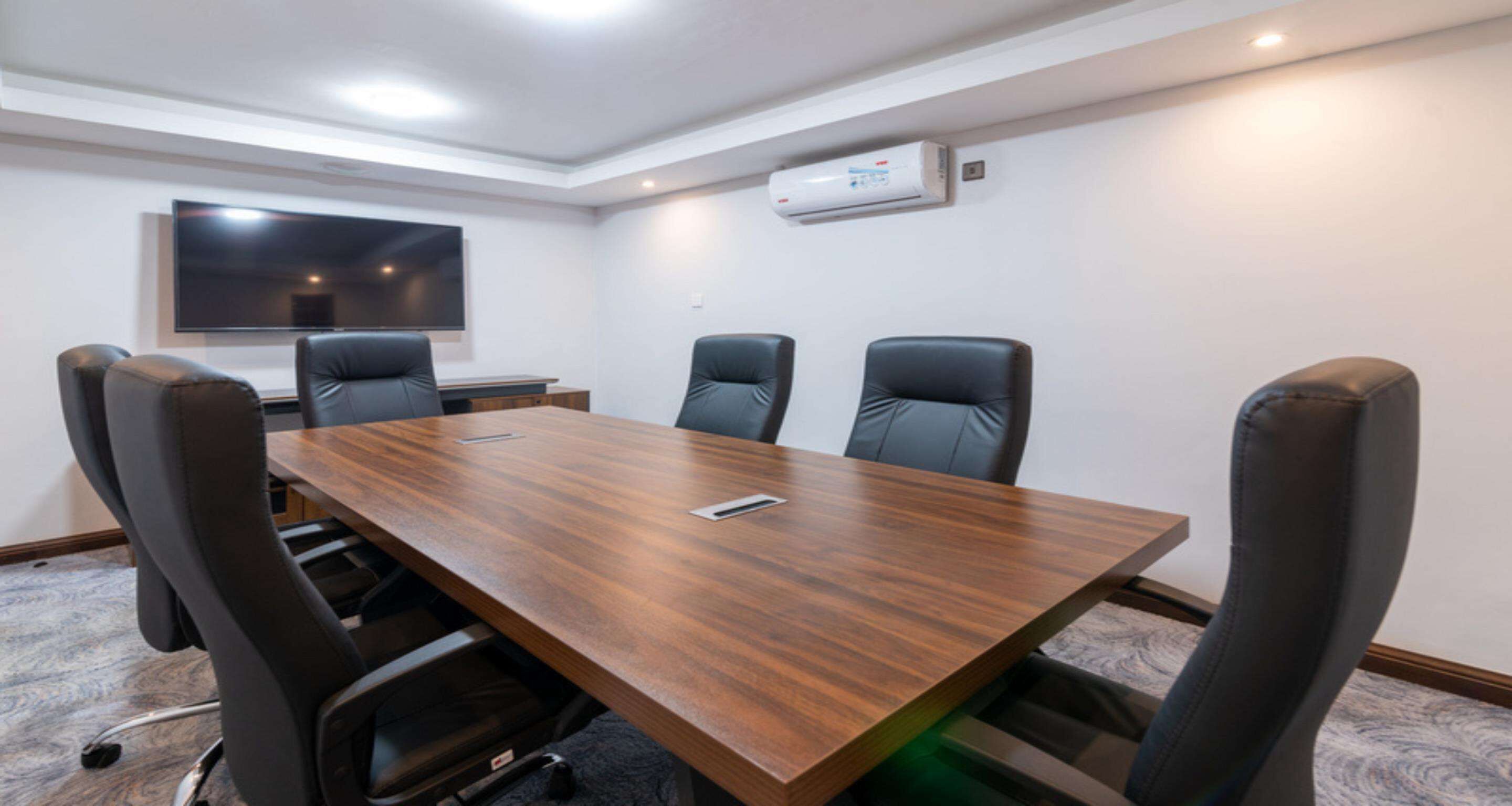 Un salón de conferencias con una larga mesa de madera, seis sillas de cuero negro, un televisor de pantalla plana en la pared y un equipo de aire acondicionado.