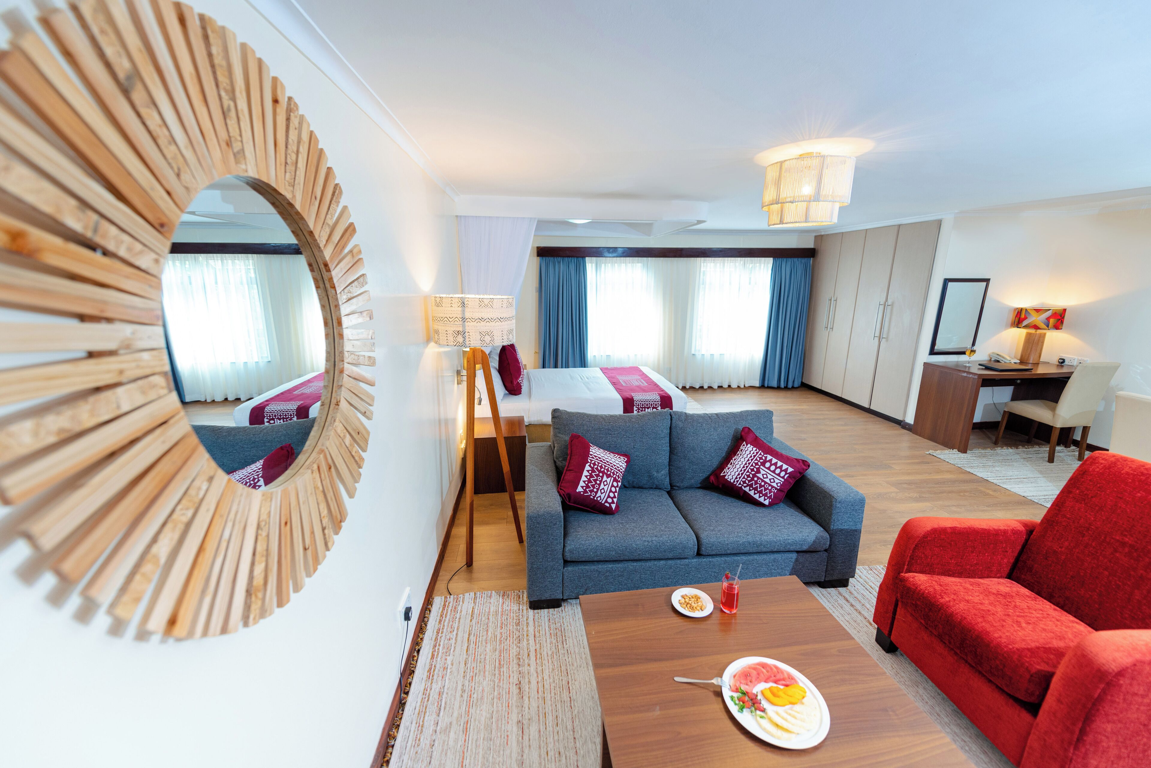 A modern hotel room with a sofa, a red armchair, a dining table with a plate of food, a mirror, and a desk with a lamp.