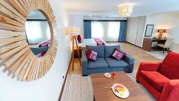 A modern hotel room with a sofa, a red armchair, a dining table with a plate of food, a mirror, and a desk with a lamp.