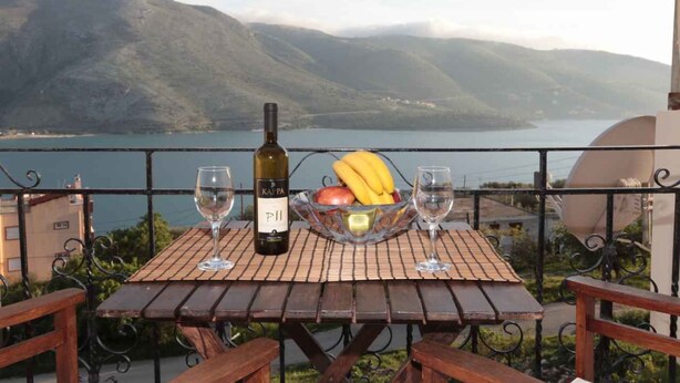 A table with a bottle of wine, a bowl of fruit, and two glasses on a balcony overlooking a lake and mountains. - Almini Apartments (Igoumenitsa)