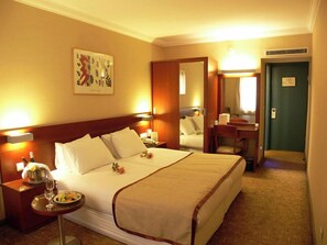 Habitación de hotel con una cama grande, mesitas de noche, un escritorio, una silla, un espejo y una puerta.