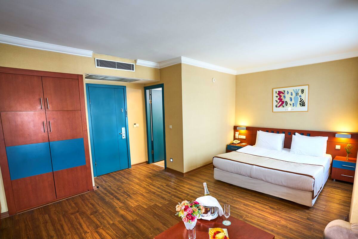 A hotel room with a bed, a nightstand, a table with a fruit platter and flowers, a wooden wardrobe, and a door.