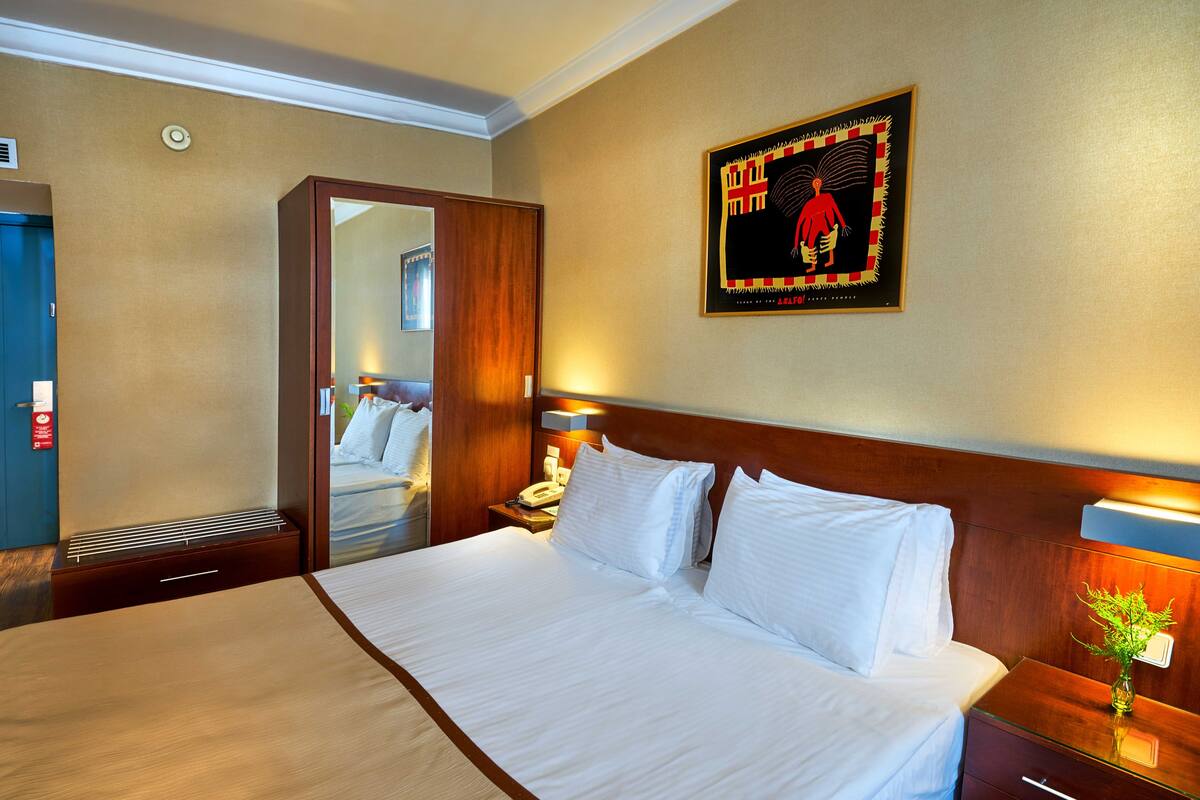 A hotel room with a large bed, a wooden headboard, a bedside table with a lamp, a mirror, and a wall-mounted artwork.