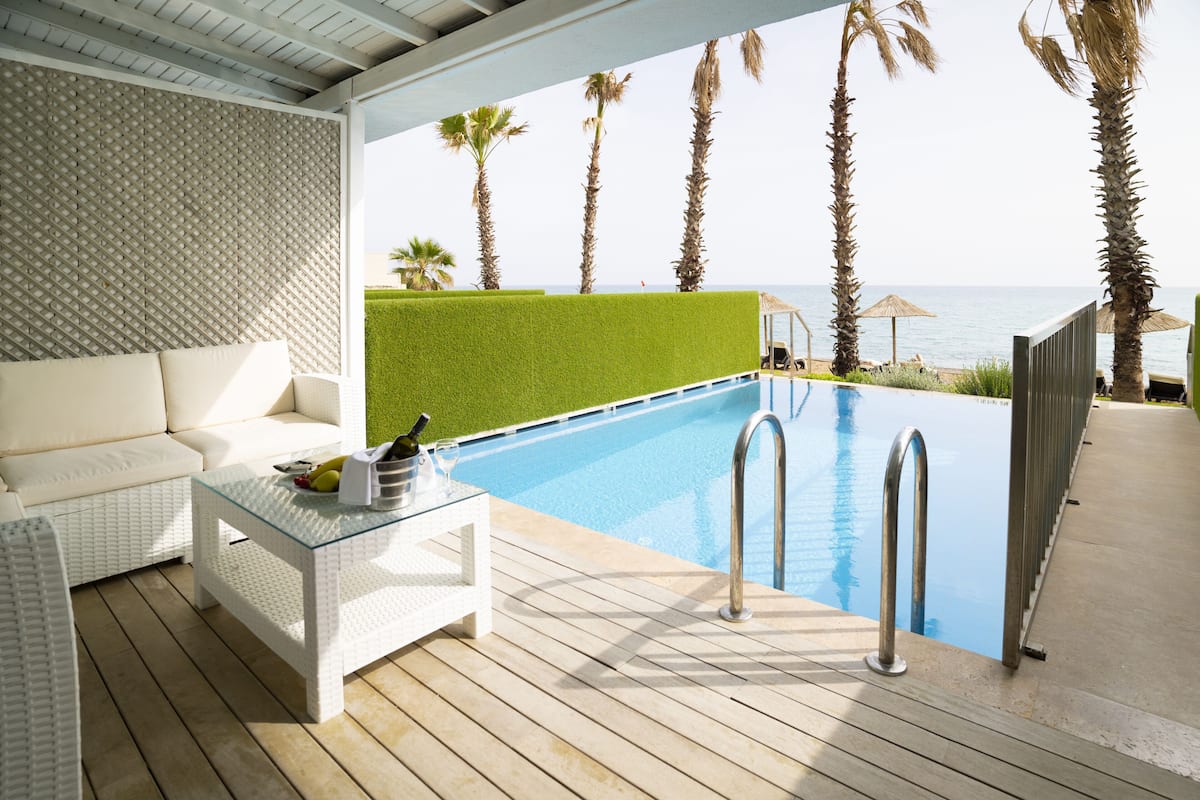 Un área junto a la piscina con un sofá blanco, una mesita de centro de vidrio y una piscina con barandilla de metal.
