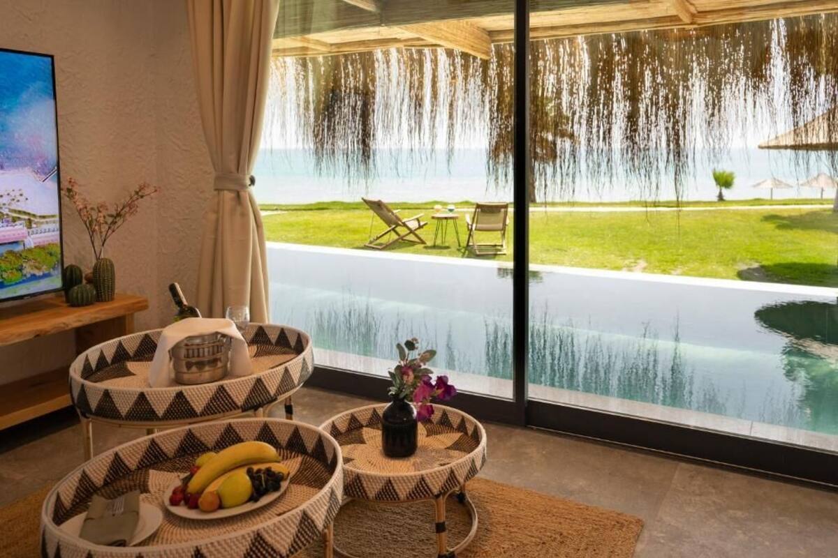 Habitación moderna con un amplio ventanal que da a una piscina y jardín. Hay dos sillas de mimbre y una mesita con un frutero.