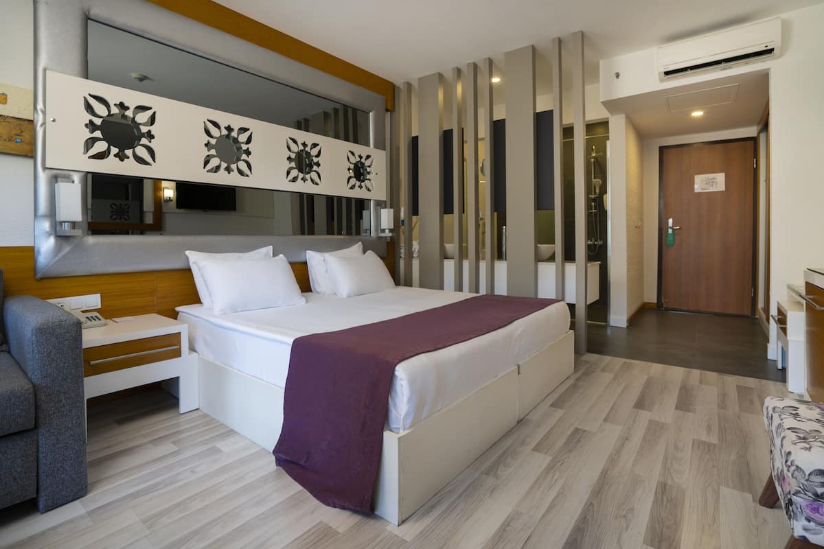 Una habitación de hotel moderna con una cama grande, un sofá gris, un cabecero de madera, un televisor y un baño visible a través de una puerta de cristal.