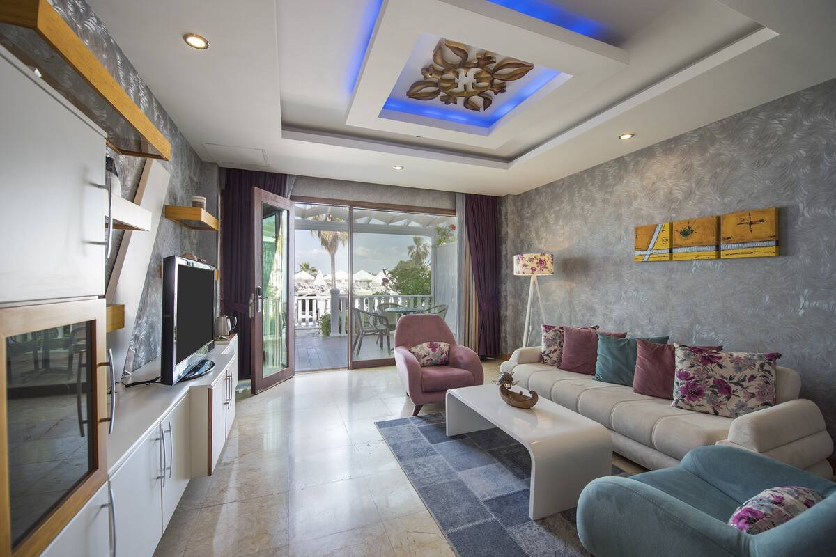Una sala moderna con un sofá, sillones, una mesa de centro y un televisor. Se ve una ventana grande con vistas a palmeras y un balcón.