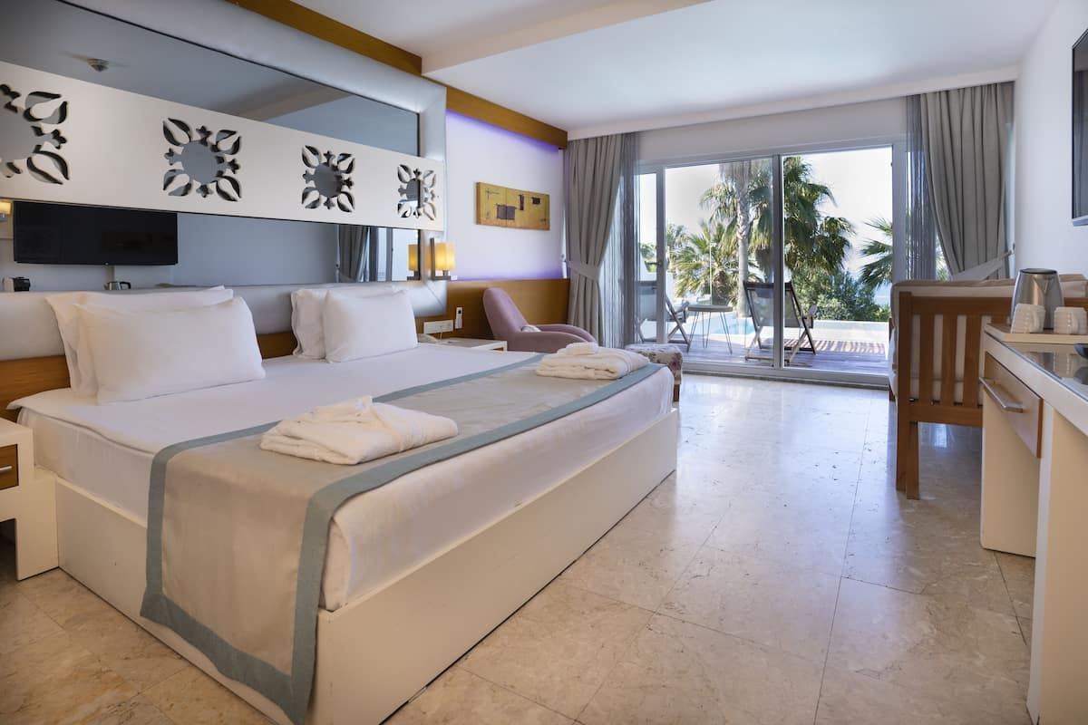 Habitación de hotel con una cama grande, un escritorio, una silla, un televisor y un balcón con vistas a palmeras.