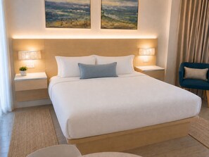 Habitación de hotel con una cama grande, dos lámparas de noche, una mesita de noche con una planta, una silla y una obra de arte en la pared.