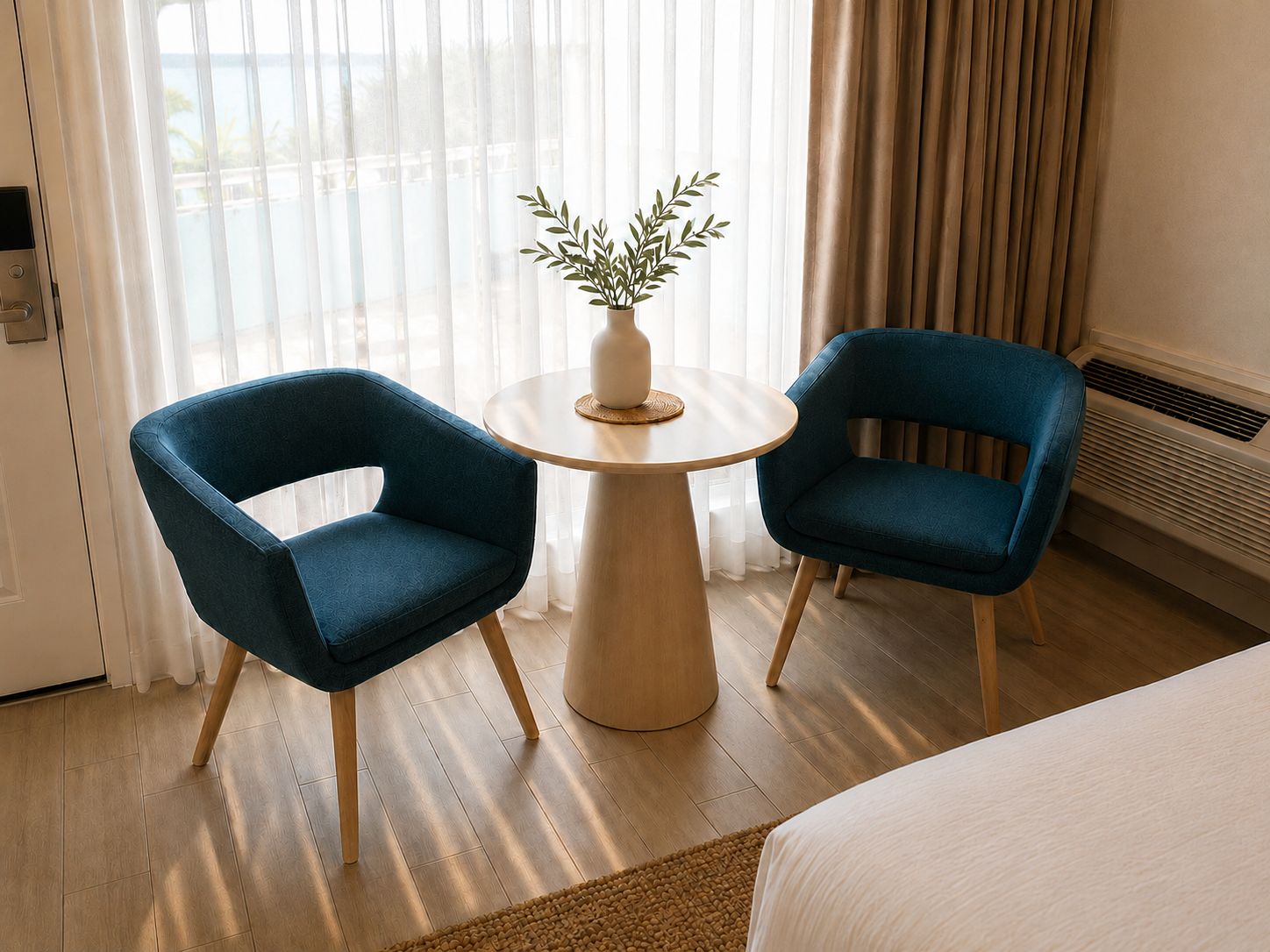 A hotel room with a bed, two blue armchairs, a small round table with a plant, and a view through sheer curtains.