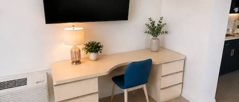 A modern, minimalist room with a flat-screen TV mounted on the wall, a desk with a lamp, a chair, and a small potted plant.