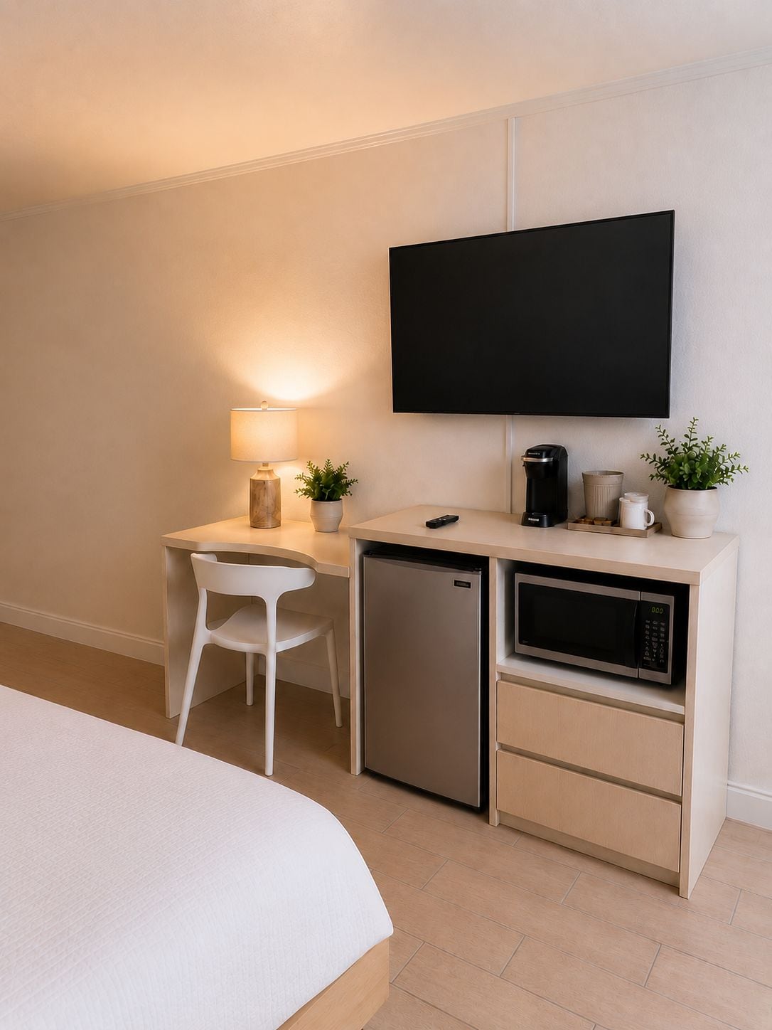 A compact hotel room with a bed, a desk with a lamp, a small refrigerator, a microwave, a coffee maker, and a flat-screen TV mounted on the wall.
