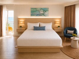 Une chambre d’hôtel avec un grand lit, deux lampes de chevet, un tableau au-dessus du lit, une chaise et une vue sur la mer depuis un balcon.