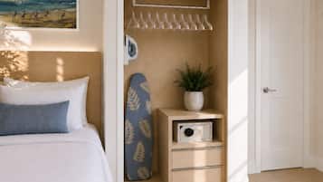 Een slaapkamer met een bed, een kast met hangende kleding, een strijkplank, een plant en een deur.