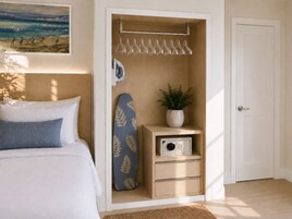 A bedroom with a bed, a closet with hanging clothes, an ironing board, a plant, and a door.