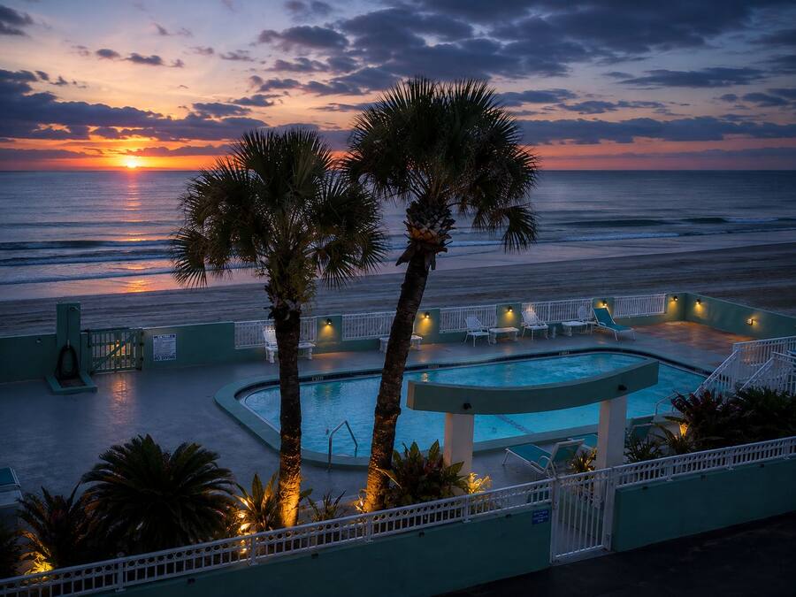 Zona de piscina con vista al océano al atardecer, palmeras y sillones de descanso.