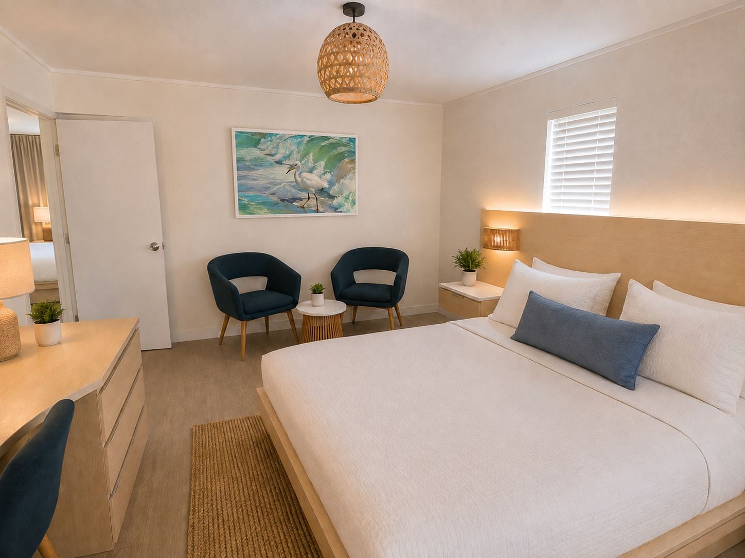 A hotel room with a bed, two chairs, a desk, a lamp, a painting, and a window with blinds.