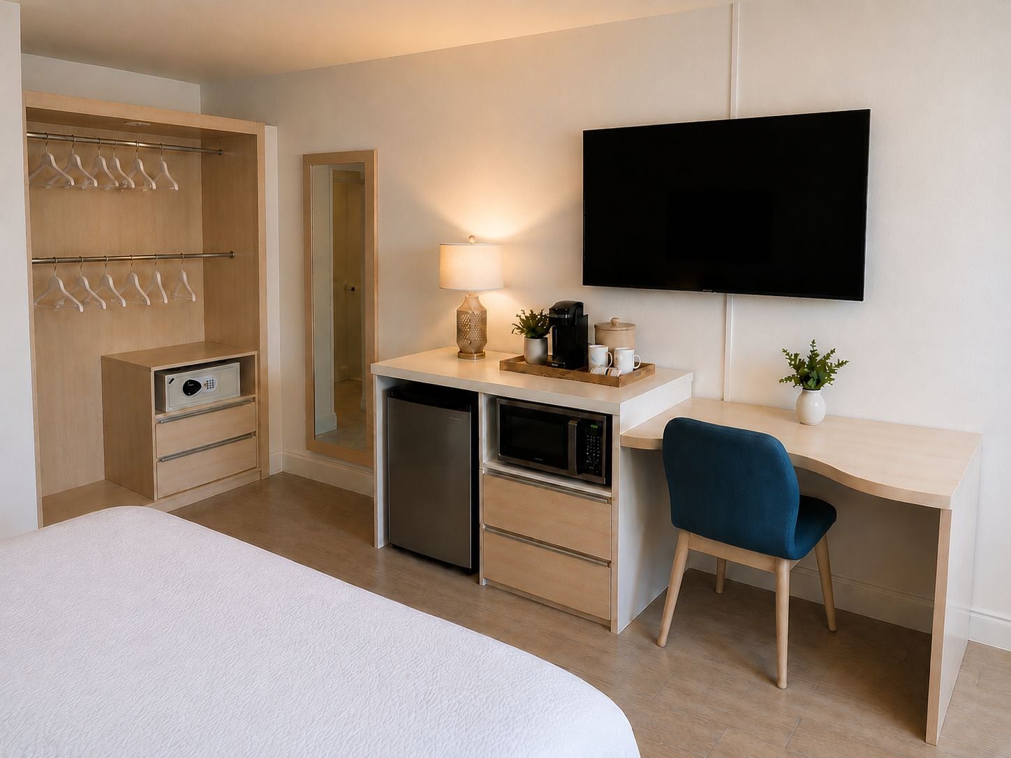 Quarto de hotel com cama, escrivaninha e cadeira, televisão, espelho, guarda-roupa e pequena cozinha.