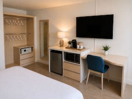 A hotel room with a bed, a desk with a chair, a television, a mirror, a wardrobe, and a small kitchenette.