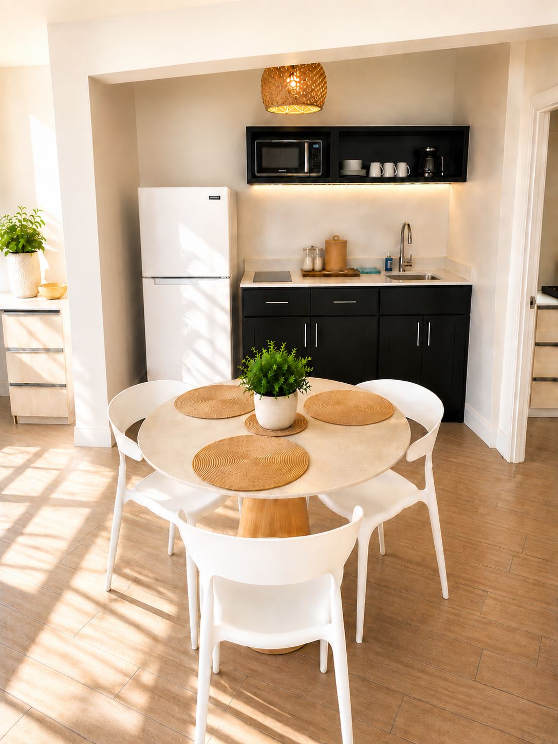A modern kitchen with a white refrigerator, black cabinets, and a microwave. There is a dining table with white chairs and a potted plant.