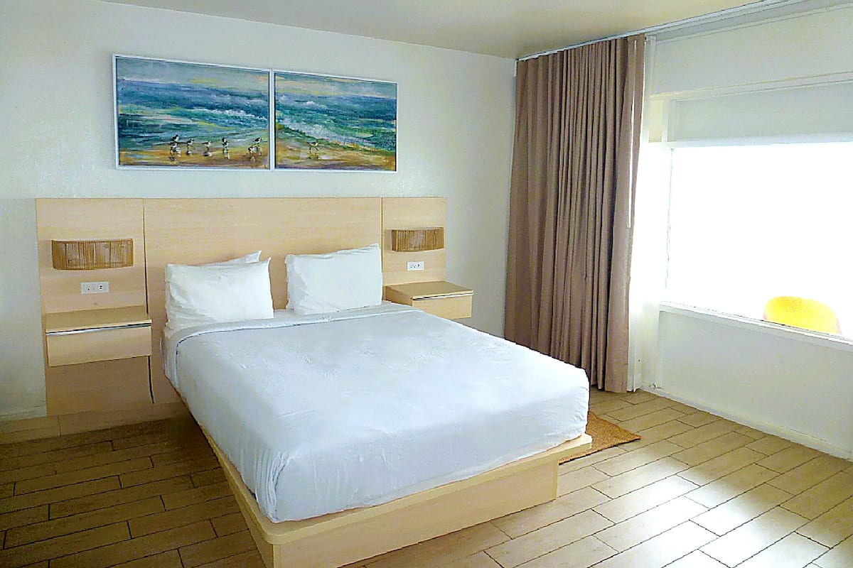 Une chambre d’hôtel avec un grand lit, deux tables de chevet, une commode, une fenêtre avec des rideaux et deux tableaux au mur.