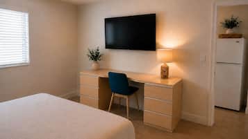 A bedroom with a bed, a desk with a chair, a television, a lamp, and a refrigerator.