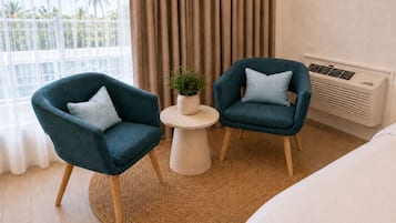 Habitación de hotel con dos sillones, una mesita con una planta, una cama y vistas a palmeras por la ventana.