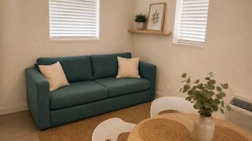 A teal sofa with two cushions, a small plant on a shelf, and a round wooden table with a vase.