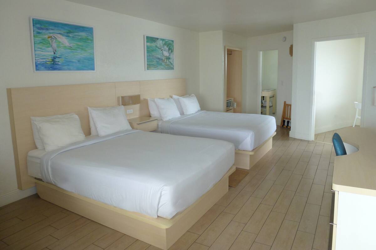 Une chambre d’hôtel avec deux lits, un bureau et une chaise. Il y a deux tableaux accrochés au mur.