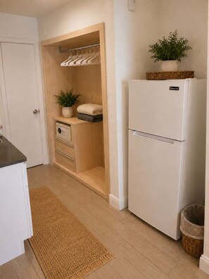Una cocina con un refrigerador blanco, gabinetes de madera, un horno empotrado, una planta pequeña y una cesta tejida encima del refrigerador.