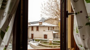 A view through a window with curtains, revealing a courtyard with a fountain, a building with a tiled roof, and a tree with bare branches. - Hotel Tre Punte (Gargnano)