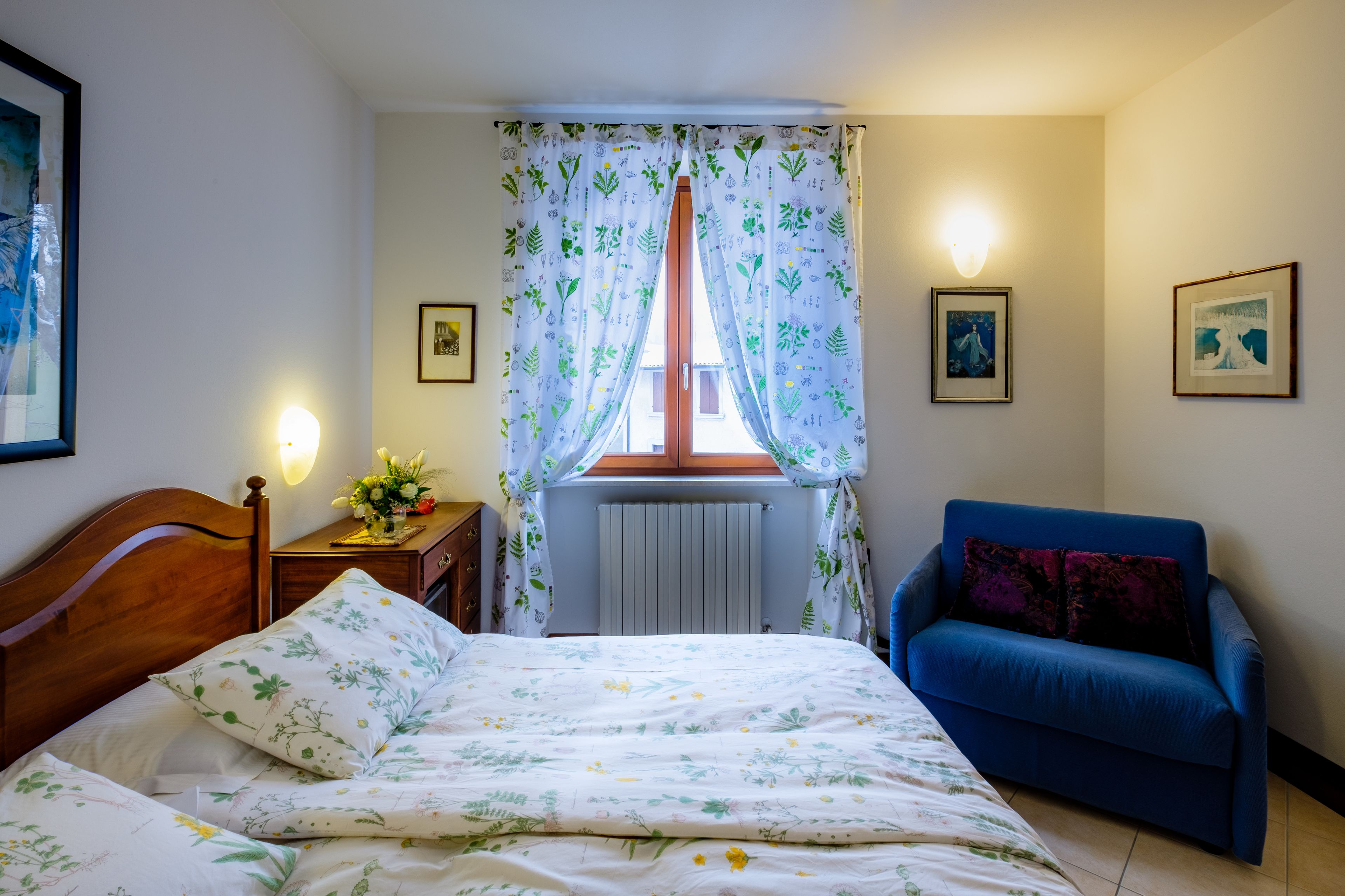 Ett sovrum med en säng, en blå soffa, ett fönster med gardiner, ett sängbord med en lampa och inramade tavlor på väggarna.