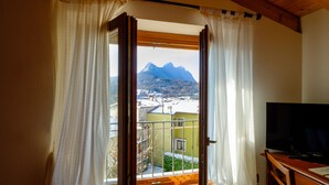 A room with a wooden ceiling, a TV on a wooden stand, and a view of mountains through an open door with curtains. - Hotel Tre Punte (Gargnano)