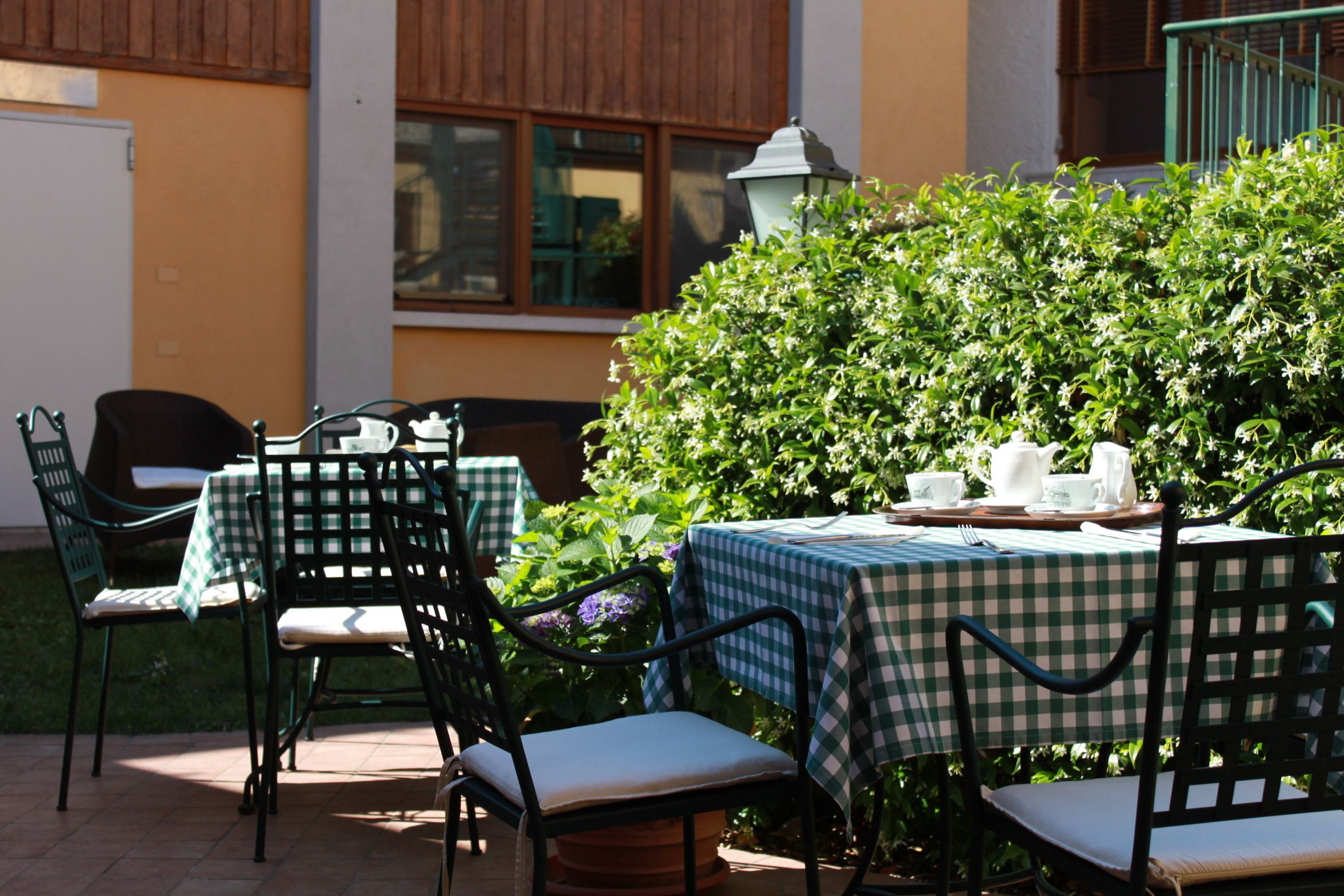An outdoor patio with a checkered tablecloth-covered table and chairs, surrounded by greenery and a building in the background.
