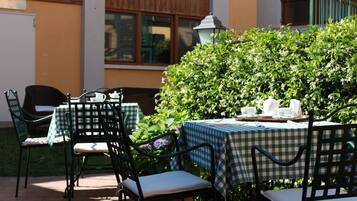 An outdoor patio with a checkered tablecloth-covered table and chairs, surrounded by greenery and a building in the background.