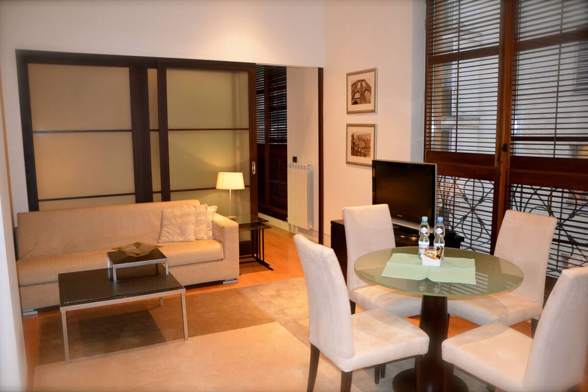 A hotel room with a sofa, a glass coffee table, a round table with chairs, a television, and a window with blinds.